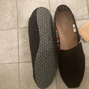 Svarta loafers från Toms  - Säljer ett par svarta TOMS från The Venice Collection. Skorna är i nyskick då skorna aldrig använts och har en färgglad randig insida. Perfekta för en avslappnad stil och passar bra till både vardag och fest. 