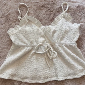 Vit linne topp med knytband - Säljer en söt vit linne topp med smala axelband och knytband i midjan. Toppen har en omlott-design och är gjord av ett texturerat material som ger en fin struktur. Perfekt för sommardagar eller en avslappnad festkväll. 🥰