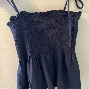 Svart ärmlös topp med volangdetaljer - Säljer en söt svart topp med smockad överdel och volangdetaljer. Den är ärmlös med tunna axelband och har en luftig passform. Perfekt för sommardagar eller en avslappnad festkväll.