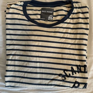 Randig t-shirt från Race Marine - Säljer en snygg randig t-shirt från Race Marine. Den är vit med mörkblå ränder och har en cool textdetalj på framsidan. T-shirten är kortärmad och perfekt för en avslappnad stil.