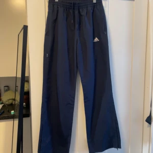 Svarta träningsbyxor från Adidas - Säljer ett par svarta träningsbyxor från Adidas. De har en bekväm elastisk midja med snörning och dragkedjor vid bensluten för justerbar passform. Perfekta för träning eller en avslappnad dag. Byxorna har också praktiska fickor med dragkedja. 