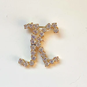 Guldfärgad brosch med strass - Säljer en superfin guldfärgad brosch formad som bokstaven K. Den är täckt med glittrande strass som verkligen fångar ljuset. Perfekt för att piffa upp en outfit till fest eller speciella tillfällen. Broschen är cirka 5 cm lång. Okänd metall 