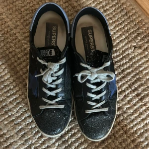 Golden goose sneakers  - Svarta Golden goose sneakers med blåa detaljer. Glitter framme vid tårna. Se bild storlek 42. Nypris ligger på strax över 5000. Mitt pris 2500kr alternativt kom med rimliga bud. 
