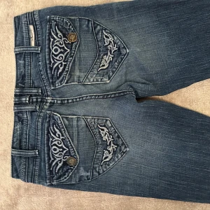 Blå jeans med broderade fickor - Säljer ett par snygga blå jeans med coola broderade detaljer på bakfickorna. De har en klassisk femficksdesign och är i en skön denimkvalitet. Perfekta för en avslappnad vardagslook!