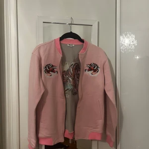 Rosa bomberjacka från Kenzo - Säljer en supercool rosa bomberjacka från Kenzo Kids. Jackan har ett snyggt tigerbroderi på både framsidan och baksidan, vilket ger den en unik och trendig look. Den är långärmad och har en dragkedja framtill. Perfekt för vår och höst!