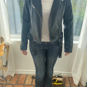 Skinnjacka - Svart skinnjacka, jackan har ett bälte som går att ta ut, jackan är svart med en dragkedja som går att dra upp ända till halsen om man lossar på knapparna, använd men inga defekter 