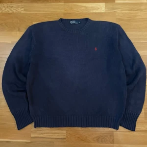 Mörkblå stickad tröja från Polo Ralph Lauren - Säljer en stilren mörkblå stickad tröja från Polo Ralph Lauren med det klassiska röda logotypbroderiet på bröstet. Tröjan har långa ärmar och ribbstickade muddar vid ärmslut och nederkant. Perfekt för kyligare dagar!