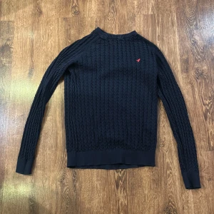 Mörkblå stickad tröja från Pier One - Säljer en snygg mörkblå stickad tröja från Pier One med ett diskret kabelstickat mönster. Tröjan har en liten röd broderad logga på bröstet och ribbade muddar vid ärmslut och nederkant. Perfekt för kyliga dagar!