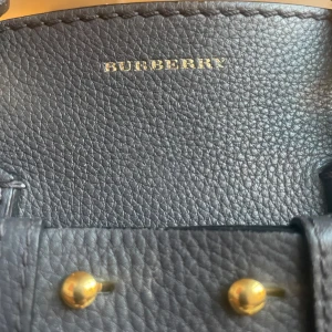 Svart skinnväska från Burberry - Elegant svart skinnväska från Burberry med stilrena detaljer i guld. Väskan har en robust design med dubbla handtag och en justerbar axelrem. Perfekt för en chic och sofistikerad look.