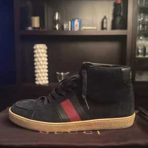 Mörkblåa sneakers från Gucci - Snygga mörkblåa sneakers från Gucci med röd randdetalj på sidan. Skorna har en beige sula och snörning framtill. Perfekta för en stilren look. Storlek 39