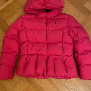 Rosa dunjacka från Polo Ralph Lauren Kids - Snygg och varm rosa dunjacka från Polo Ralph Lauren Kids i L men oxå xs/s  i vuxen storlek. Jackan har en huva och är perfekt för kyliga dagar. Den är quiltad och har en klassisk design med dragkedja framtill.