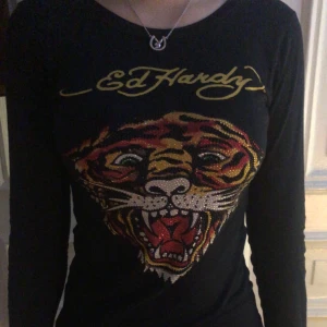 Svart långärmad tröja från Ed Hardy - Cool svart långärmad tröja från Ed Hardy med ett stort tigertryck i färgglada nyanser på framsidan och Ed Hardy-logga i guld på både fram- och baksidan. Perfekt för en edgy look!