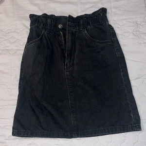 Svart jeanskjol med resårmidja - Snygg svart jeanskjol med resår i midjan och knapp- och dragkedjestängning framtill. Kjolen har praktiska fickor både fram och bak, vilket ger en avslappnad look. Perfekt för en casual outfit.