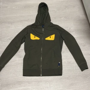 Fendi kofta - Snygg grön hoodie med dragkedja och huva. Framsidan har unika gula ögonliknande detaljer som ger en cool look. Perfekt för en avslappnad stil. Man får även en vara som kostar 3000kr vid köpet av Fendi koftan 