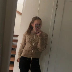 Beige stickad tröja med volangdetaljer - Säljer en mysig beige stickad tröja med volangdetaljer. Tröjan har långa ärmar och en hög krage, perfekt för kyligare dagar. Passar bra till både jeans och kjol.