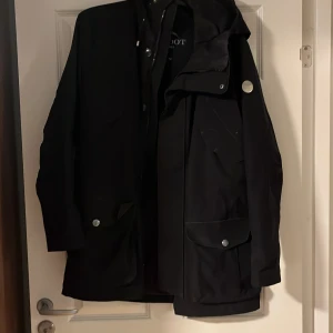 Svart jacka från Snoot storlek medium - Snygg svart jacka från Snoot med huva och praktiska fickor. Jackan har en stilren design med knappar och dragkedja framtill. Perfekt för kyligare dagar.