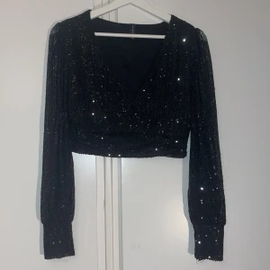 Svart glittrig topp från Amisu - Snygg svart glittrig topp från Amisu med långa ärmar och en croppad passform. Perfekt till nyår med sitt glittrande material och stilrena design. Toppen har en v-ringad framsida och ribbade muddar vid ärmsluten.