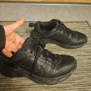Svarta Nike sneakers - Säljer ett par svarta Nike sneakers med snörning. Skorna har en robust design med Nike-loggan på sidan och är perfekta för vardagsbruk. De är tillverkade i syntetmaterial och har en bekväm passform. Enda skavanken är insidan av hälen men märks ej av.