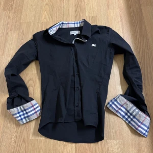 Svart skjorta från Burberry - Snygg svart skjorta från Burberry med klassiskt rutigt mönster på insidan av kragen och ärmsluten. Passform är 165-170! Hör av dig om du har frågor eller funderingar! Priset kan diskuteras vid snabb affär!🙌🤝 Såklart ÄKTA!