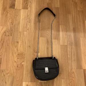 Svart axelväska med kedja - Snygg svart axelväska i krokodilmönstrat material med silverfärgad kedja och låsdetalj. Perfekt för att ge en stilren touch till din outfit.