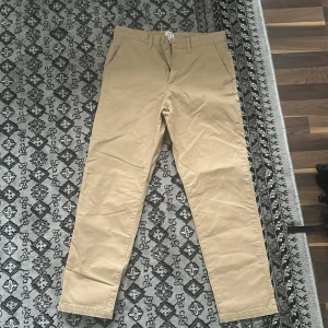 Beige chinos från H&M - Snygga beige chinos från H&M med normal passform. Byxorna har knappar och bälteshällor, vilket gör dem perfekta för en stilren look. De är tillverkade i ett bekvämt bomullsmaterial.