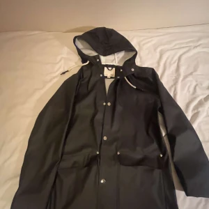 Svart regnjacka från Gellstad - Snygg svart regnjacka från Gellstad med huva och knappar framtill. Jackan har en stilren design med ljus insida och praktiska fickor. Perfekt för regniga dagar.