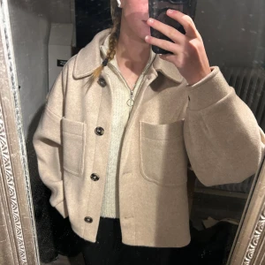 Vår/höstjacka från NA-KD - Säljer en stilren beige jacka från NA-KD. Jackan har en klassisk krage och knäppning med stora knappar framtill. Den är croppad och har två stora fickor på bröstet. Perfekt för en trendig look! Jackan är lite ”luftigare/pösigare” 