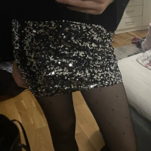 Svart glitterkjol  - Snygg svart kortkjol täckt med glittrande paljetter. Perfekt för att sticka ut på festen! Kjolen har en tight passform och är ett måste för garderoben.