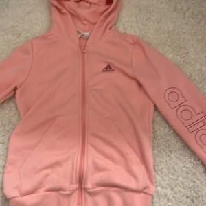 Rosa hoodie från Adidas - Säljer en snygg rosa hoodie från Adidas med dragkedja och huva. Den har långa ärmar och det står adidas på vänstra armen. Perfekt för en avslappnad stil.