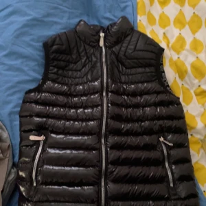 Svart dunväst från Philipp Plein - Snygg svart dunväst från Philipp Plein med glansig finish och dragkedja framtill. Västen har två fickor med dragkedjor och ett stilrent mönster på insidan. Perfekt för kyligare dagar. Finns även qrcode på insidan så man kommer till deras hemsida, 