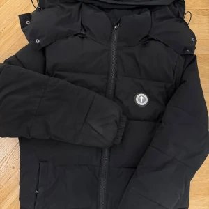 Svart pufferjacka från Trapstar - Snygg svart pufferjacka från Trapstar med dragkedja och huva i storlek M den sitter bra och har används ett tag. Pris kan diskuteras vid snabb affär ( OBS. En liten defekt på dragkedjan där den har gått sönder )