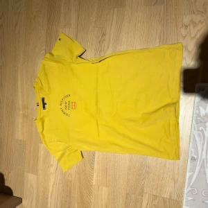Gul t-shirt från Tommy Hilfiger - Snygg gul t-shirt från Tommy Hilfiger med tryck 'New York' på bröstet. Den har korta ärmar och är tillverkad i mjuk bomull. Perfekt för en avslappnad stil.