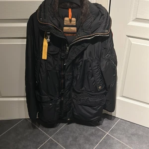 Svart parkajacka från Parajumpers - Snygg svart parkajacka från Parajumpers med flera praktiska fickor och en cool gul detalj vid dragkedjan. Jackan har en hög krage och är perfekt för kyligare väder.