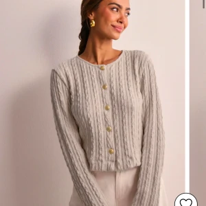 Beige ribbad kofta med knappar - Säljer en stilren beige kofta med ribbad struktur och dekorativa knappar i guld. Koftan har en croppad passform och långa ärmar, perfekt för lager på lager-stil. Ett mångsidigt plagg för både vardag och fest. Oanvänd