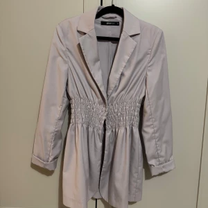 Ljuslila kavaj från Ginatricot - Elegant beige kavaj med smockdetaljer i midjan som ger en snygg siluett. Kavajen har långa ärmar och klassisk krage, perfekt för en stilren look.