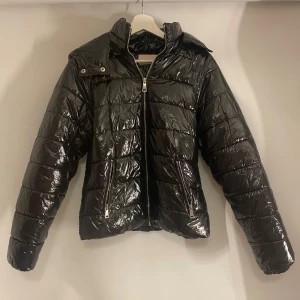 Svart glansig pufferjacka - Snygg svart glansig pufferjacka med dragkedja och huva. Perfekt för kyliga dagar med sin quiltade design som håller värmen. Jackan har långa ärmar och en stilren look.