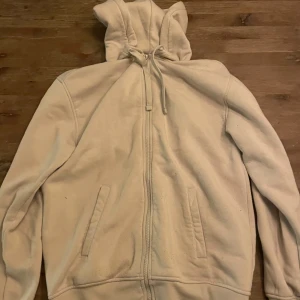 Beige hoodie med dragkedja - Säljer en beige hoodie med dragkedja och huva. Tröjan har långa ärmar och ribbade muddar vid ärmslut och nederkant. Perfekt för en avslappnad stil.