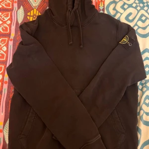 Svart hoodie från Stone Island - Säljer en svart hoodie från Stone Island med klassisk logga på ärmen. Tröjan har en hög krage med dragsko och en stor ficka framtill. Perfekt för en stilren och bekväm look.