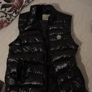 Svart dunväst från Moncler - Snygg svart dunväst från Moncler med glansig finish och dragkedja framtill. Västen har en broderad logotyp på bröstet och praktiska fickor.