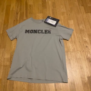Grå t-shirt från Moncler - Snygg grå t-shirt från Moncler med deras ikoniska logga tryckt på bröstet. Perfekt för en stilren och avslappnad look. T-shirten är kortärmad och har en normal passform.