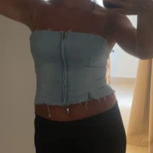 Ljusblå tubtopp i jeans - Trendig ljusblå tubtopp i jeansmaterial med råa kanter och dragkedja framtill. Perfekt för en avslappnad sommarlook. Den är med mening lite sliten, men det är för att man ska få ”vintage” looken!