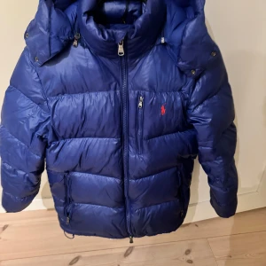 Blå dunjacka från Polo Ralph Lauren - Säljer en snygg blå dunjacka från Polo Ralph Lauren. Jackan har en dragkedja framtill och en praktisk huva. Perfekt för kyliga dagar med sin pufferdesign som håller dig varm och bekväm. Jackan är i ett gott skick och är självklart äkta. Om fler bilder önskas är det bara och be mig skicka😃Pris kan möjligtvis diskuteras.  Skriv om du har några funderingar MVH.