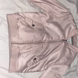 Rosa bomberjacka från Zadig & Voltaire - Snygg rosa bomberjacka från Zadig & Voltaire med dragkedja och broderad text på ryggen. Jackan har långa ärmar och ribbade muddar. Perfekt för en cool och avslappnad stil. Jackan är perfekt för både vår och höst knappast använd. Jackan är även för yngre barn från kanske 6-8