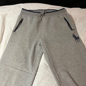Grå mjukisbyxor från Polo Ralph Lauren - Snygga grå mjukisbyxor från Polo Ralph Lauren med mörkblå detaljer och dragsko i midjan. Byxorna har två fickor med dragkedja och en broderad logotyp på benet. Endast använda ett fåtal gånger. Storlek 18-20 år ungdom