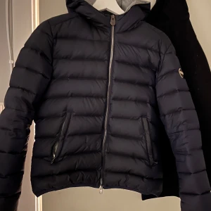 Svart dunjacka från Colmar - Snygg svart dunjacka från Colmar med huva och dragkedja. Jackan har en quiltad design och två praktiska fickor med dragkedjor. Perfekt för kyligare dagar. Den har också ett igensytt hål på framsidan men inget som syns eller som man tänker på!