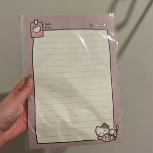 Sött anteckningsblock med kattmotiv - Ett gulligt anteckningsblock med lila kant och en söt kattillustration i hörnet. Perfekt för att skriva ner datum och titlar. Blocket har linjerade sidor och är inslaget i plast.