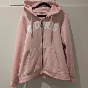 Rosa tröja med dragkedja från Björn Borg - Säljer en rosa tröja från Björn Borg med dragkedja och stor text på bröstet. Den har en mysig huva med dragsko och praktiska fickor framtill. Perfekt för en avslappnad stil. Begagnat skick, finns spår av användning. Se t.ex svart prick på sista bilden.