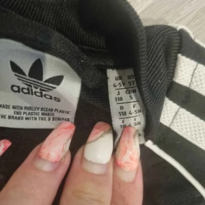 Svart träningsset från Adidas - Snyggt svart träningsset från Adidas med de klassiska tre vita ränderna längs ärmar och ben. Jackan har dragkedja och byxorna har elastisk midja för bekväm passform. Perfekt för sport och fritid.  OBS BARN DRESS