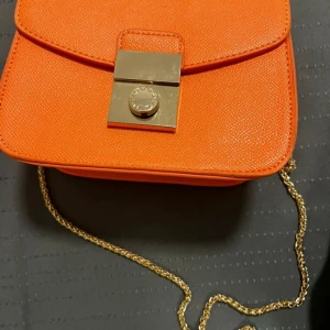 Orange axelväska med kedja - Snygg orange axelväska med guldfärgad kedja och låsdetalj. Perfekt för att ge en färgklick till din outfit. Väskan har en praktisk storlek och ett stilrent utseende.