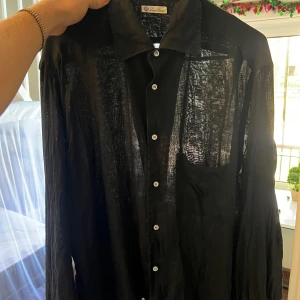 Svart skjorta från Loro Piana - Snygg svart skjorta från Loro Piana i ett lätt och luftigt material (linnen). Skjortan har långa ärmar och knappar framtill. Perfekt för en stilren look.           Storlek: S skulle jag säga,                                              Finns två hål på framsidan, men dom är väldigt små. Syns på bild. 